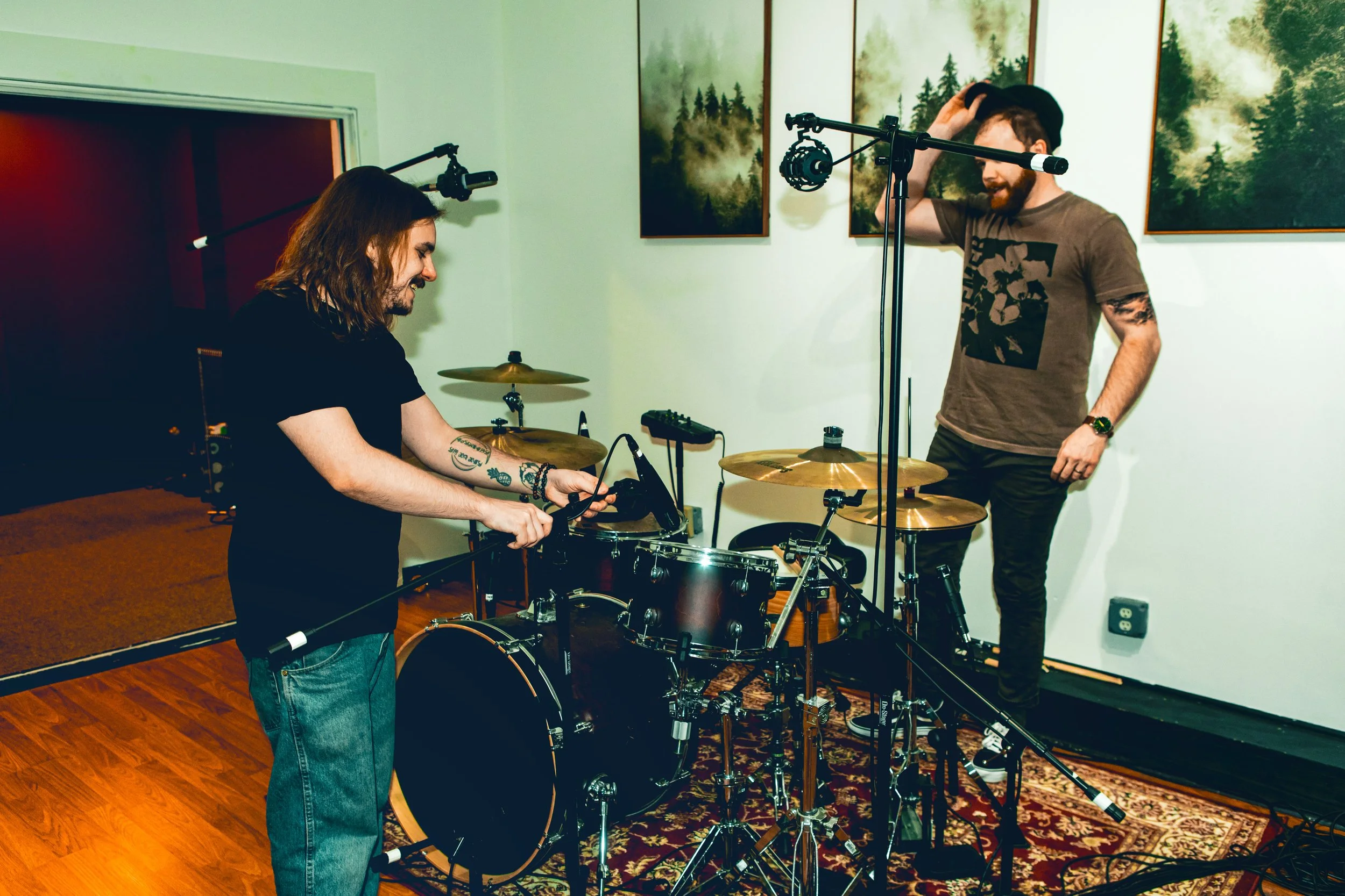 Two musicians in a recording studio, one playing drums and the other adjusting headphones, with forest-themed artwork on the walls.