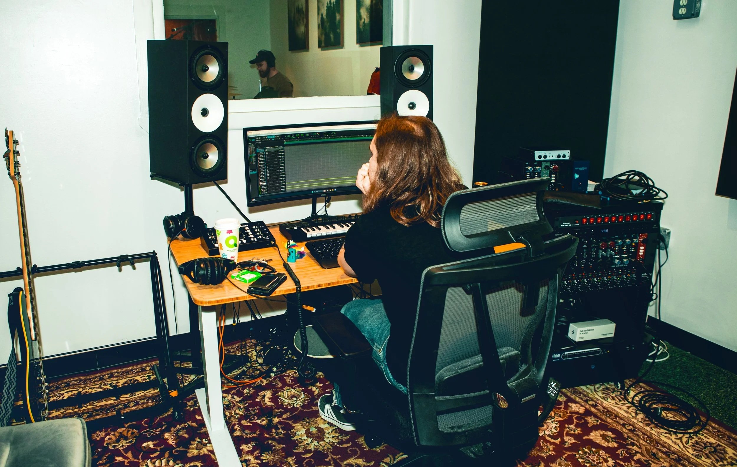 A music producer working at a computer in a recording studio, with large studio monitors, audio equipment, and headphones on the desk.