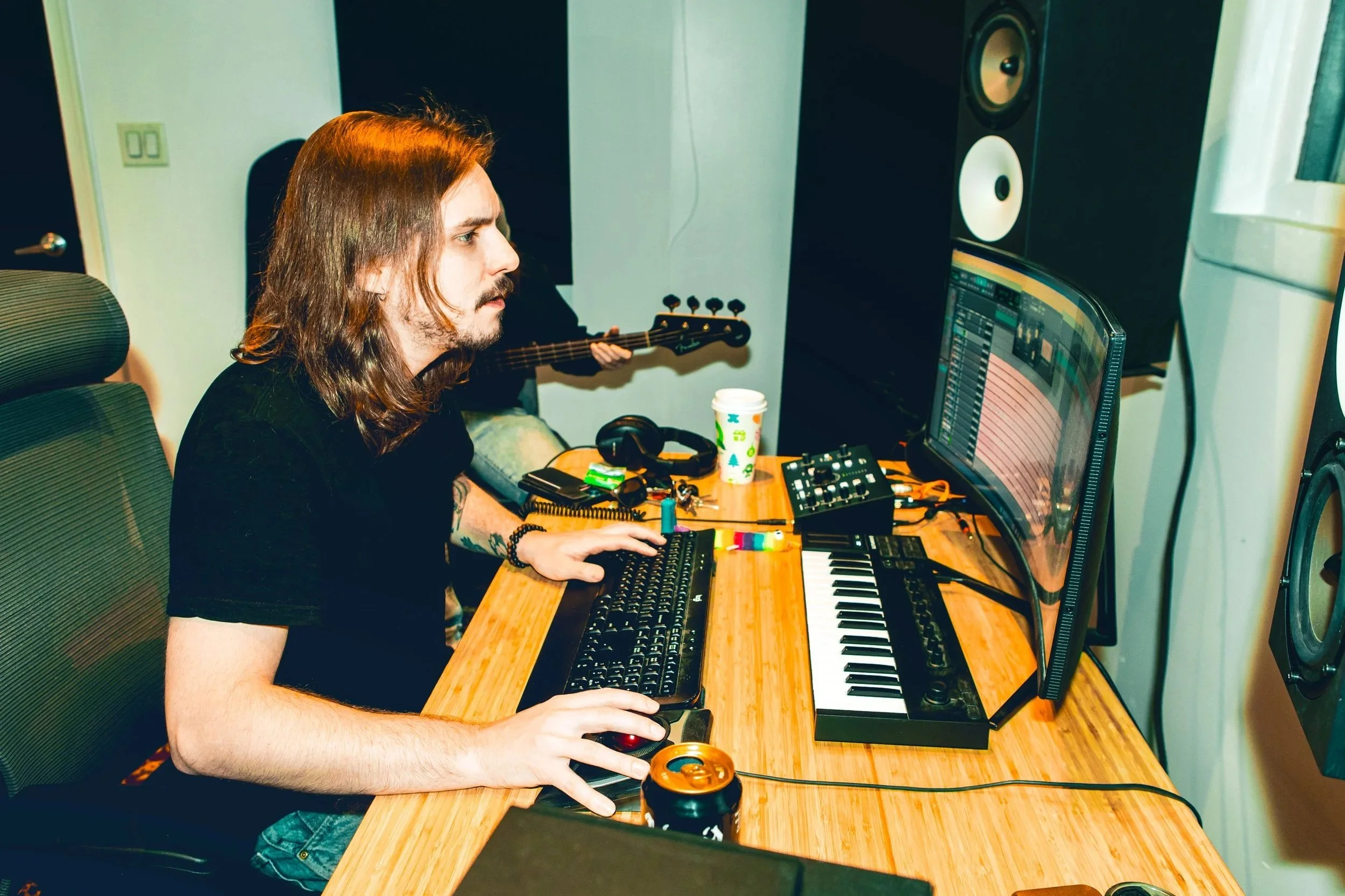 A man with long hair and a beard wearing a black t-shirt working on music production at a cluttered desk with a computer, keyboard, headphones, and a beverage can, in a recording studio with speakers on the wall.