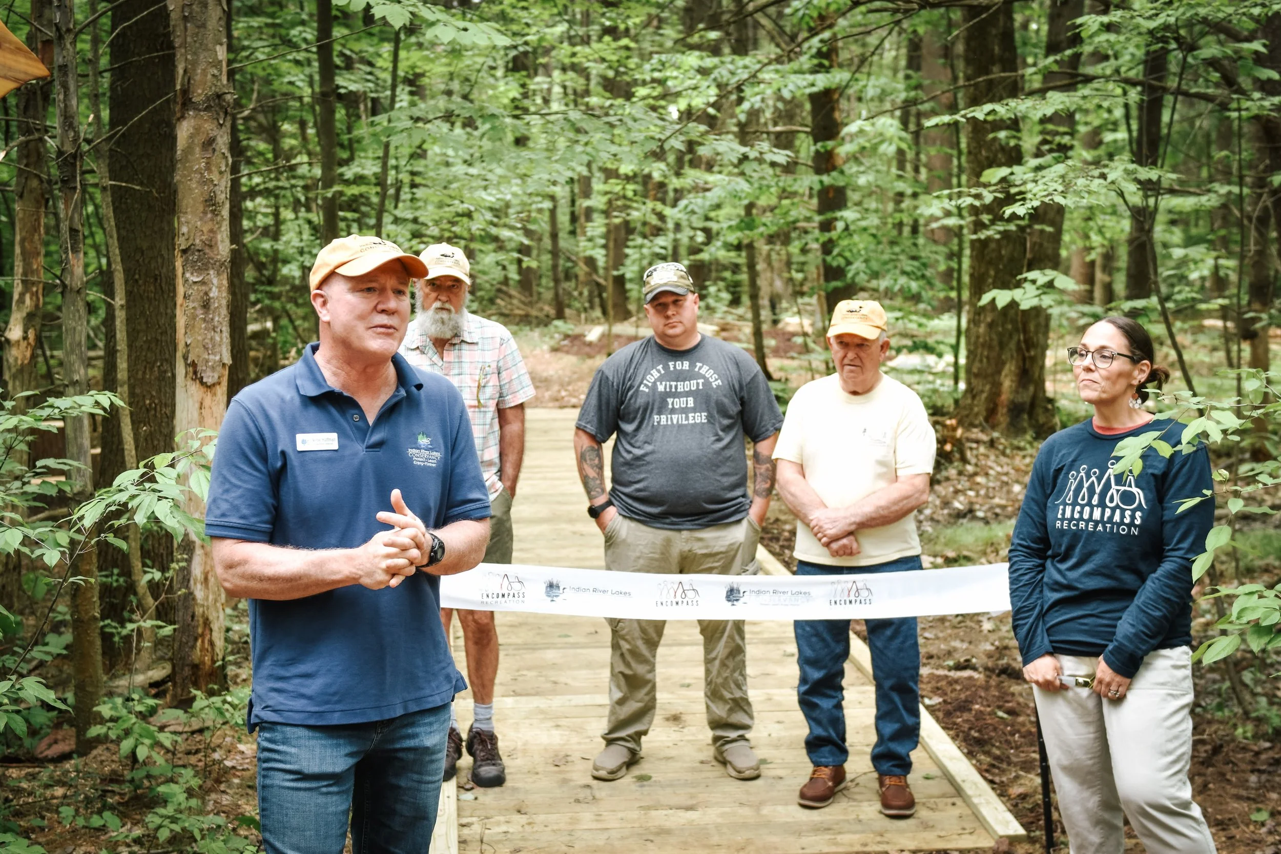 Group of five people standing on a wooden trail in a forest during a ribbon-cutting event for Encompass Recreation, a nonprofit in Northern NY providing inclusive recreational opportunities for youth with higher support needs
