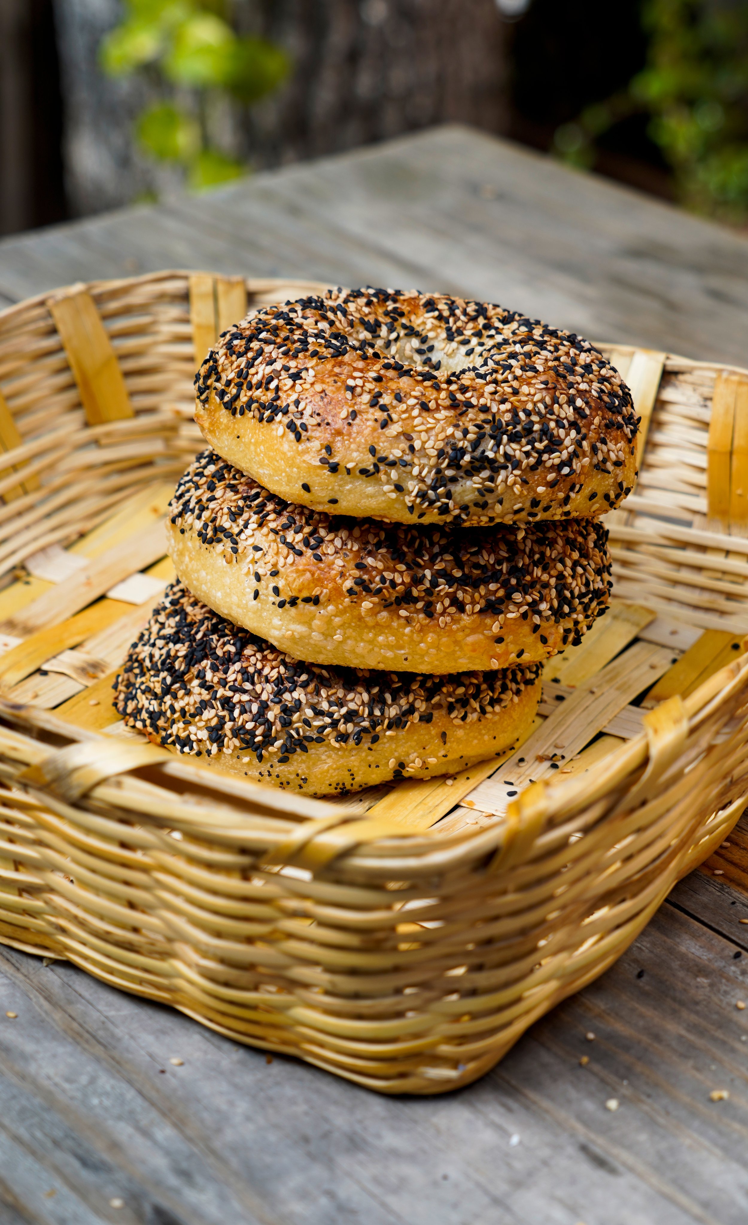 Tres bagels en una cesta de mimbre sobre una mesa de madera, cubiertos con semillas de sésamo negro y blanco.
