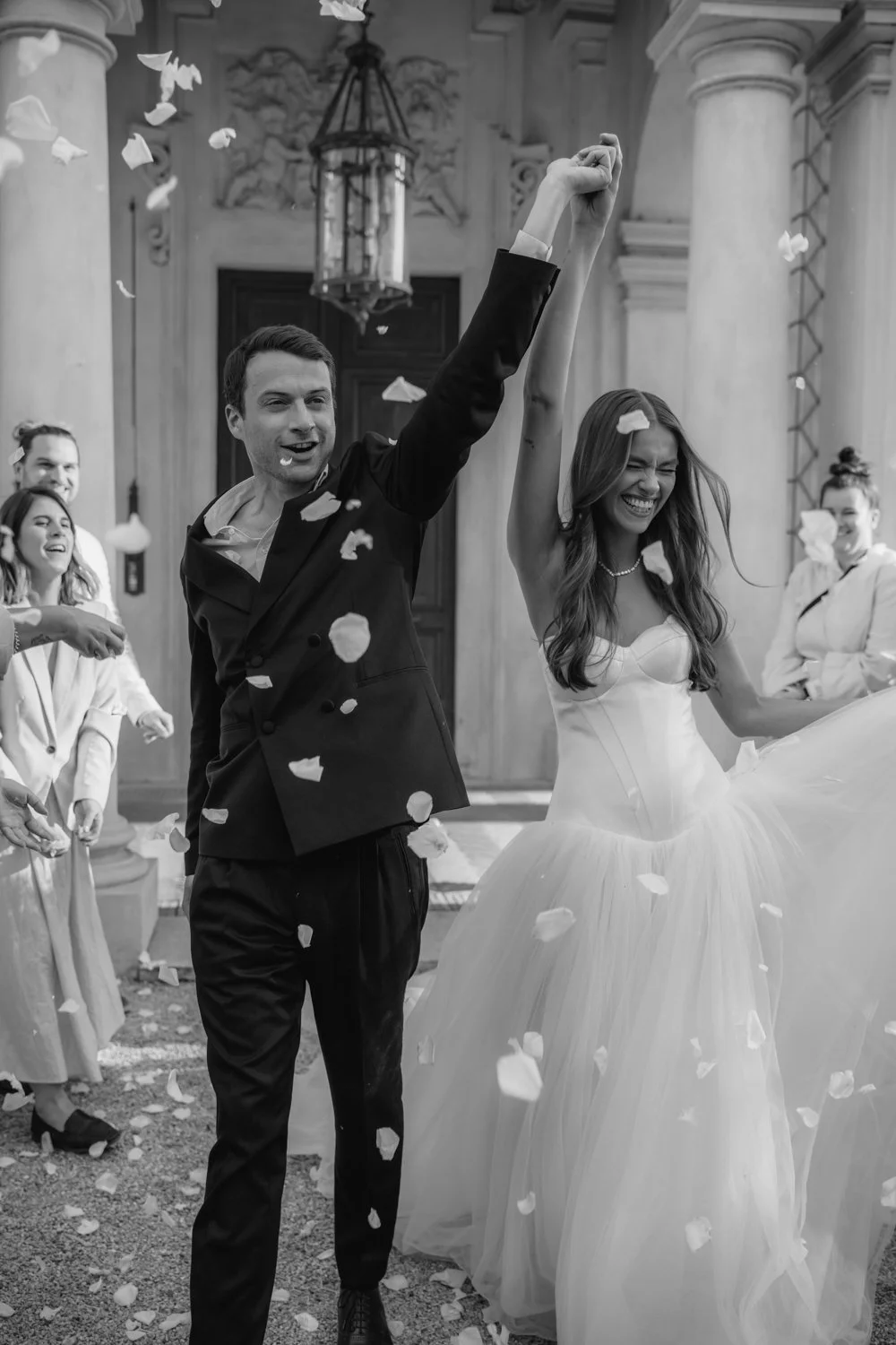 A bride and groom celebrating their wedding day outside a grand building, surrounded by friends throwing white flower petals, smiling and cheering.