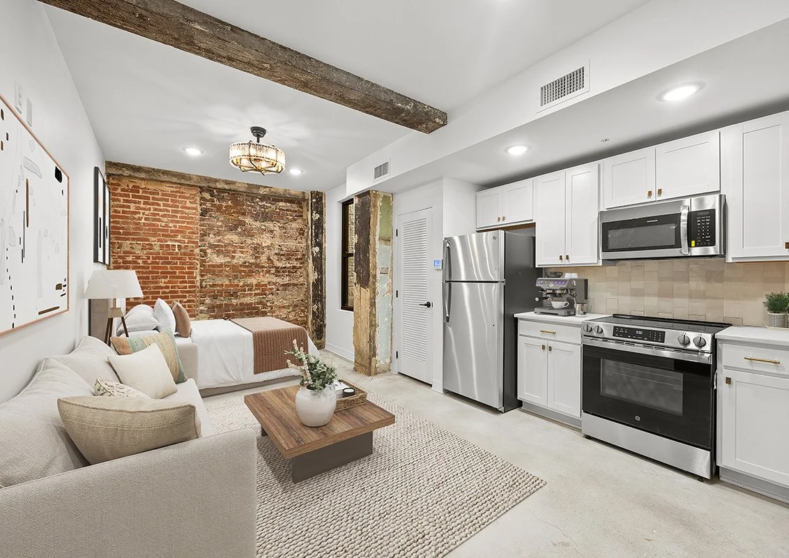 Open-concept living space with a white sofa and pillows, a wooden coffee table with a potted plant, an exposed brick wall, and modern kitchen appliances including a refrigerator, microwave, oven, and coffee maker.