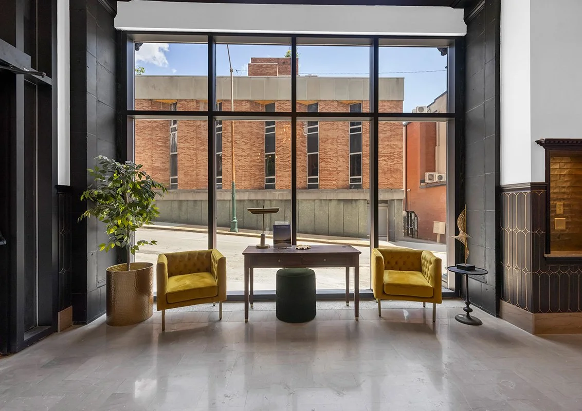 Interior view of a modern lobby or seating area with large floor-to-ceiling windows, two yellow armchairs, a wooden table with decorative items, a potted plant, and city buildings visible outside.
