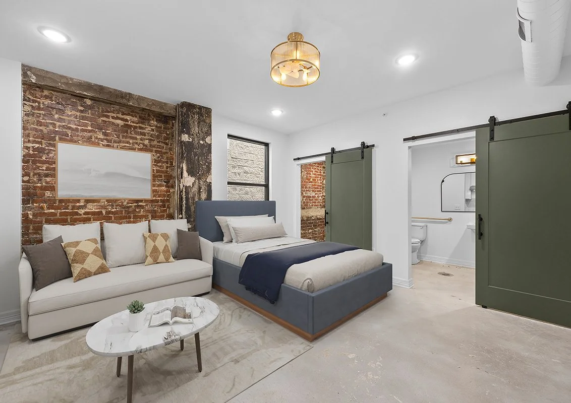 A modern bedroom with an exposed brick wall, a gray bed, a white sofa with pillows, and a small oval marble coffee table. There are two green sliding barn doors leading to a bathroom with a toilet and a window. The ceiling has recessed lighting and a