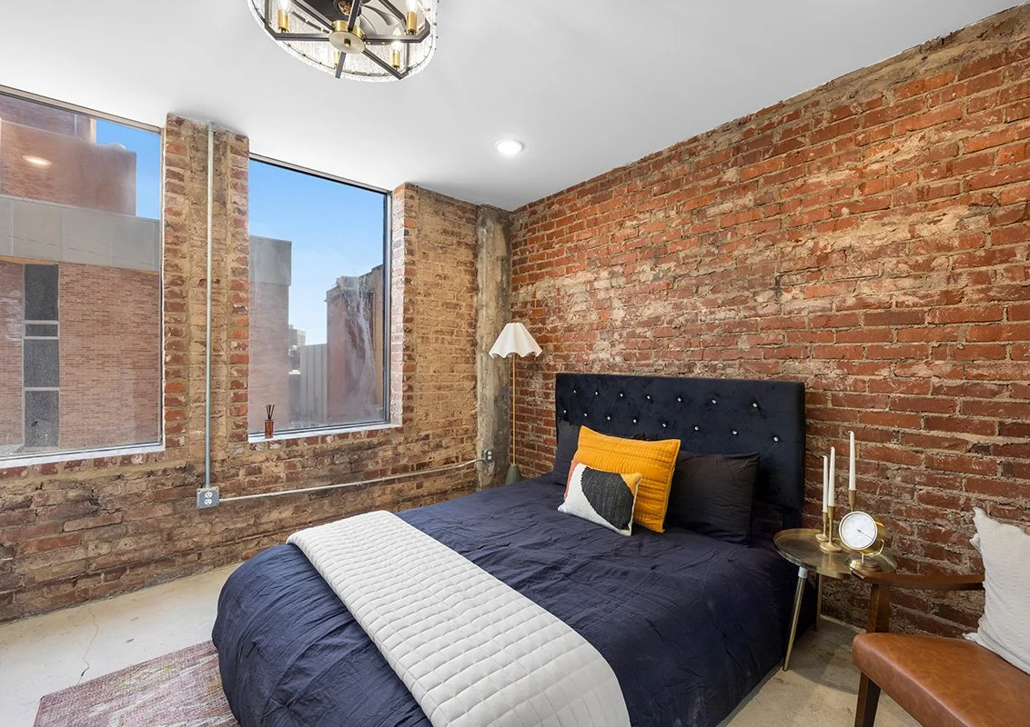 Bedroom with exposed brick walls, large windows, a black upholstered bed with orange and pastel pillows, a white quilted blanket, a round gold side table with white candles and a clock, a white floor lamp, and a light fixture on the ceiling.