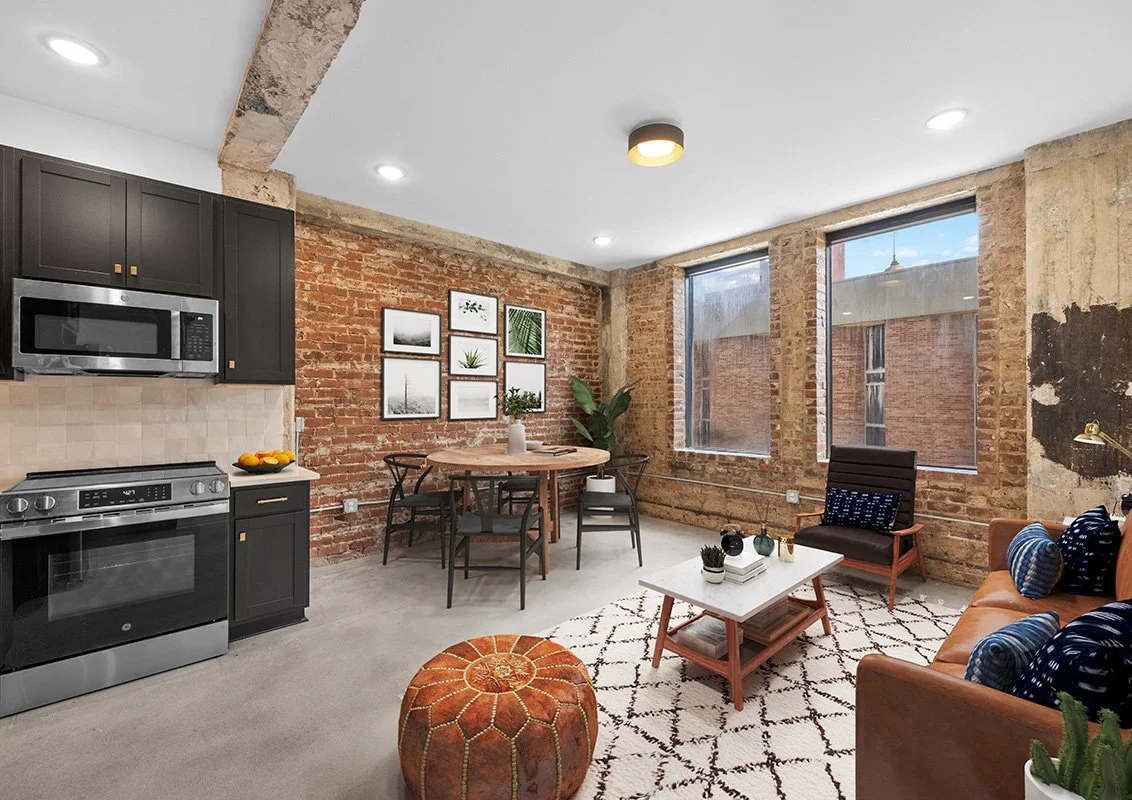 Living room with exposed brick wall, black cabinetry, kitchen appliances, dining table, armchair, sofa with blue pillows, coffee table, and large windows.