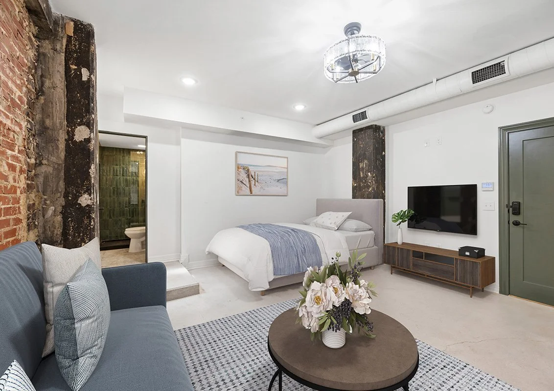 Modern bedroom with a bed, gray headboard, TV, wooden TV stand, blue sofa, and floral centerpiece on a round coffee table. The room has exposed brick and concrete walls, a chandelier, and a doorway leading to a bathroom.