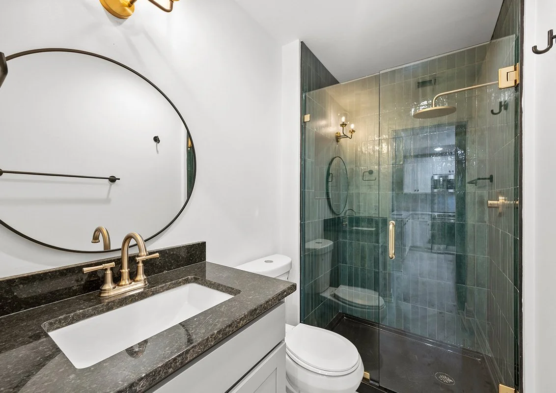 Bathroom with white walls, black granite vanity with an oval mirror, gold fixtures, a toilet, and a glass-enclosed shower with dark green tiles and gold fixtures.