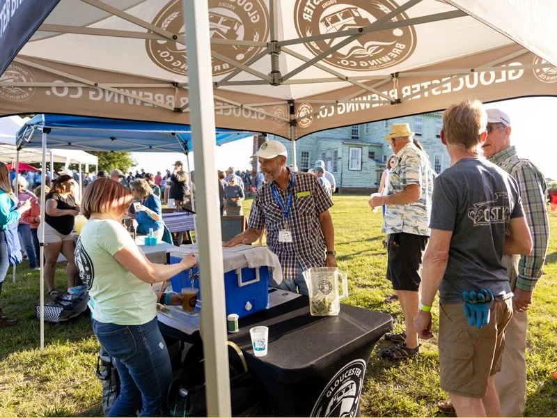 People gathered at an outdoor event under tents, with one person serving food or drinks at a booth, and others socializing on a grassy area.