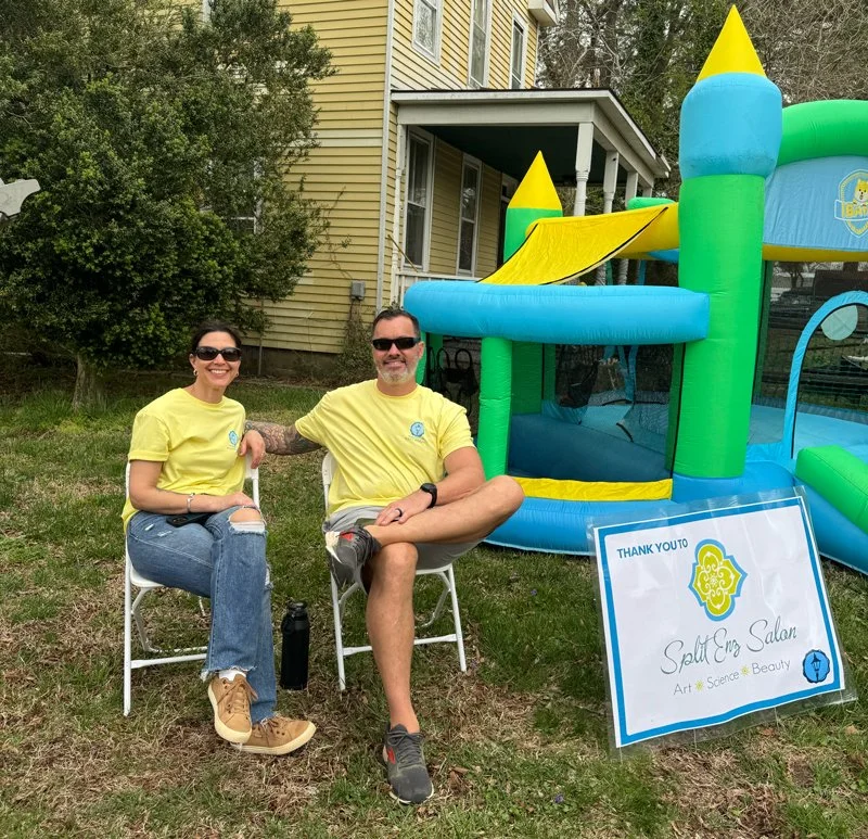 Two people wearing yellow shirts sitting on chairs outdoors in front of an inflatable bounce house, with a sign for 'Speak Easy Salon' nearby.
