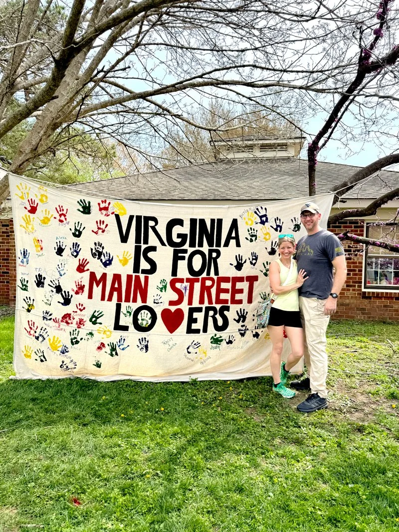 A couple standing in front of a large banner that reads "Virginia is for Main Street Lovers." The banner is decorated with colorful handprints and signatures. The couple is smiling and posing for the photo, with the trees and a brick house in the background.