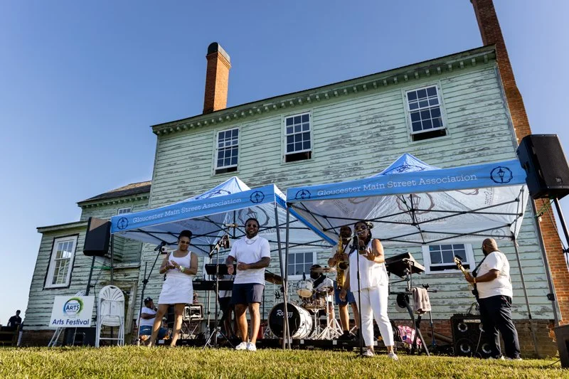 Outdoor music band performing under tents at an arts festival in front of a weathered, two-story house.