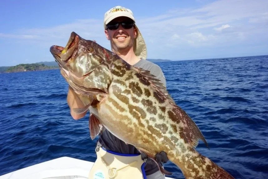 Guest holding a large grouper aboard Fish Tale 2 fishing charter with Guanacaste coastline in the background