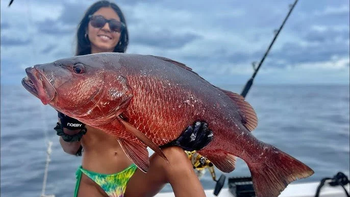 Woman holding a large red fish on a boat with water and fishing rod in the background.