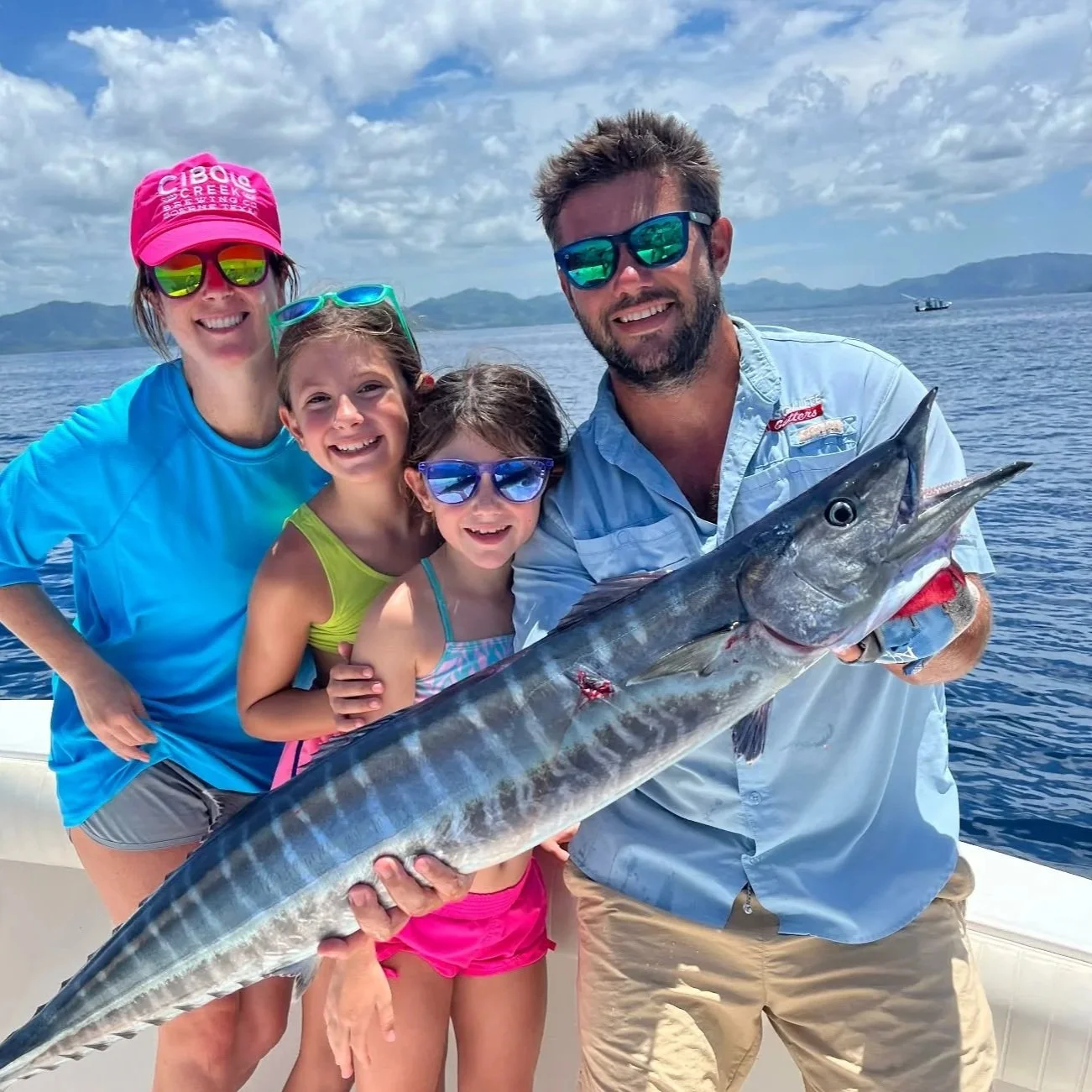 A family of four, including two young girls and their parents, smiling and holding a large fish on a boat in the ocean under a partly cloudy sky.