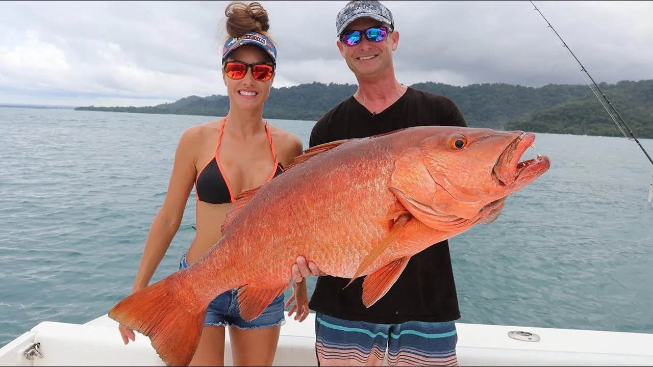 A woman and a man holding a large orange fish on a boat with water and hills in the background.