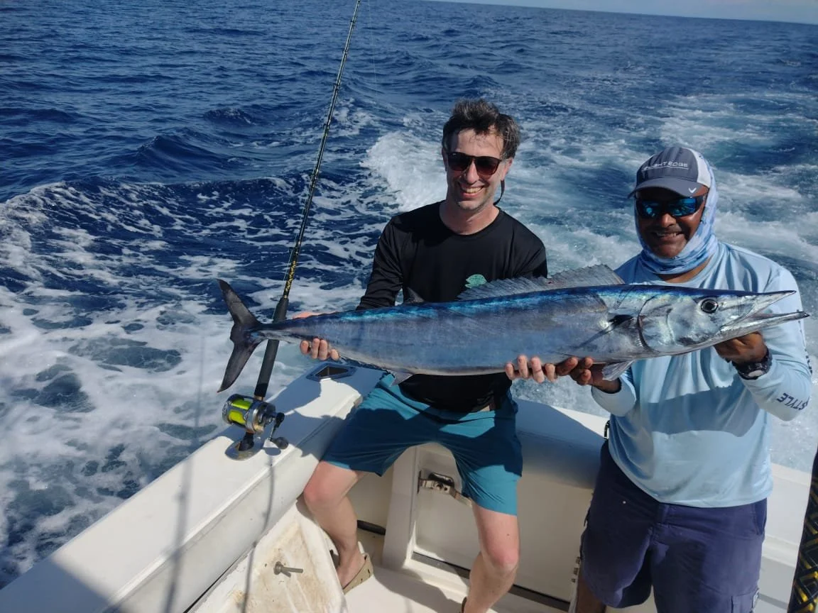 Two men on a boat holding a large fish, smiling, with water and wake in the background.