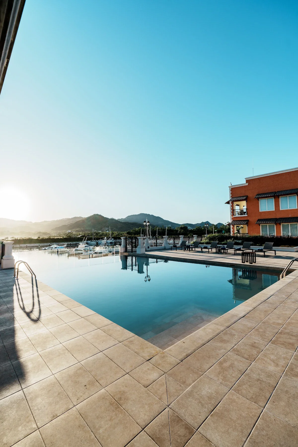 An outdoor swimming pool area with lounge chairs, a railing, and a view of sailboats docked in the Marina Flamingo, located in Playa Flamingo, Guanacaste, Costa Rica.