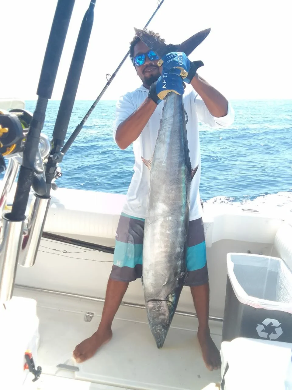 A man holding a large fish on a boat at sea, wearing sunglasses, gloves, and casual clothing.
