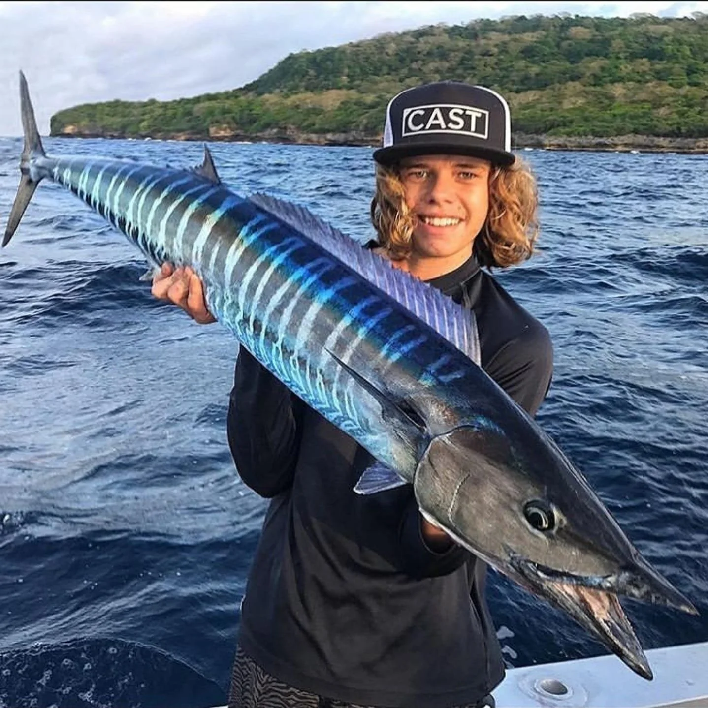 A young man holding a large, colorful fish on a boat in the ocean with an island in the background.