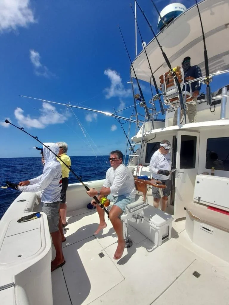 People fishing off the side of a white yacht on the open ocean under a bright blue sky with a few clouds.