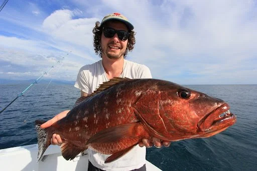 Man wearing sunglasses and a hat holding a large fish on a boat in the water with a cloudy sky in the background.