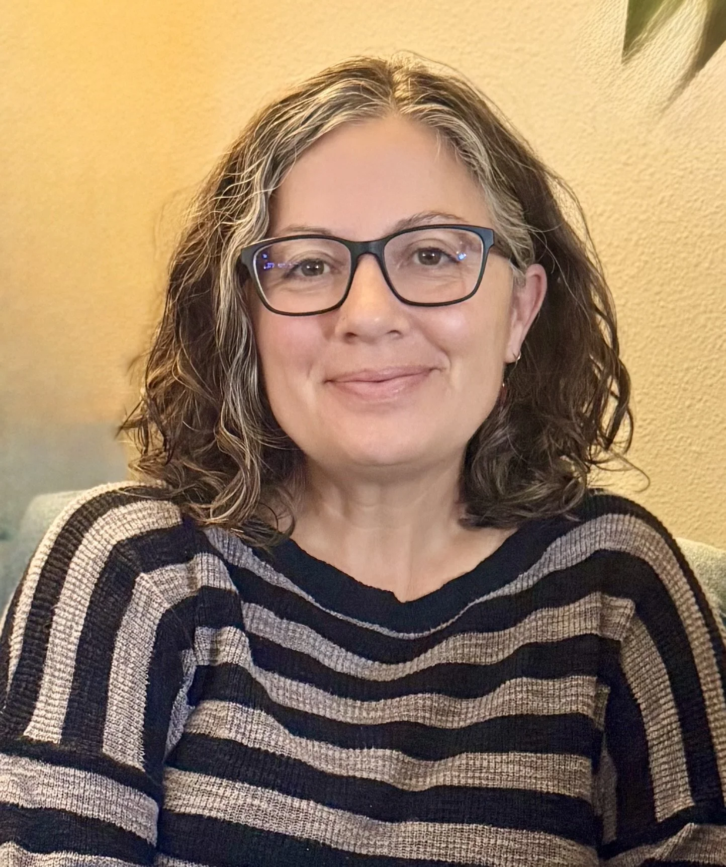 A woman with curly, shoulder-length hair and glasses, wearing a striped sweater, smiling softly in front of a beige wall.