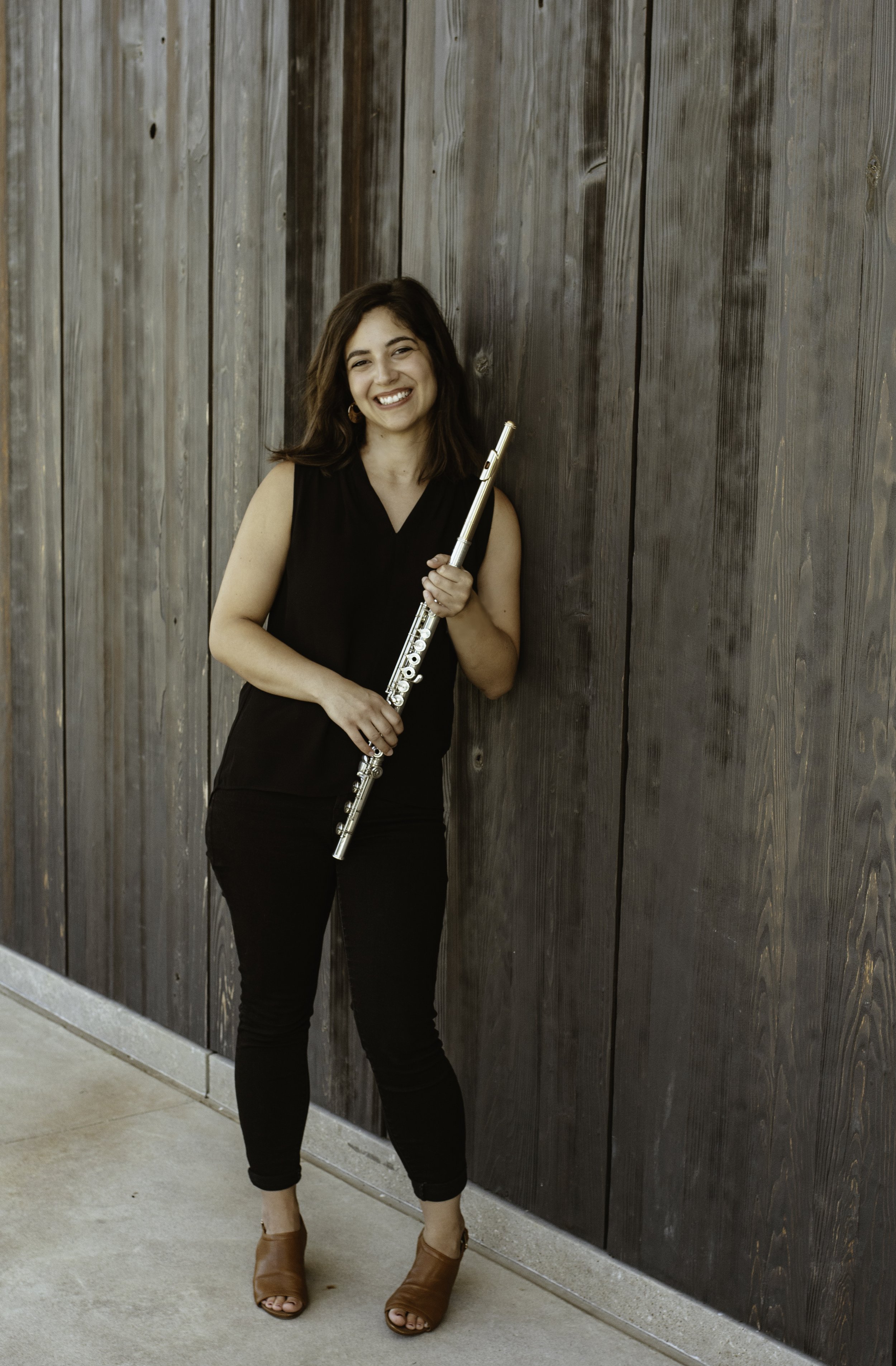 A woman with shoulder-length dark hair and light skin, dressed in a sleeveless black top and black pants, holds a silver flute while standing against a dark wooden wall, smiling at the camera.