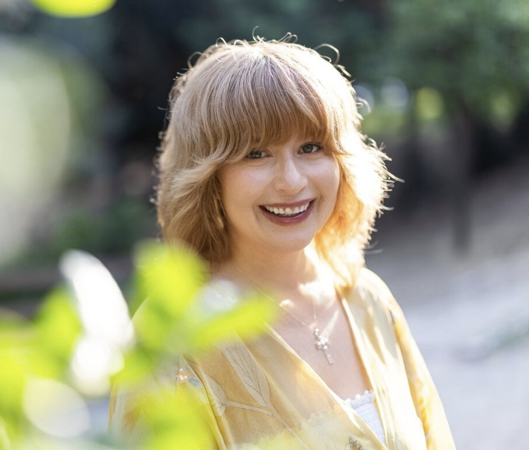 A smiling woman with red hair and a yellow blouse outdoors with greenery and sunlight in the background.
