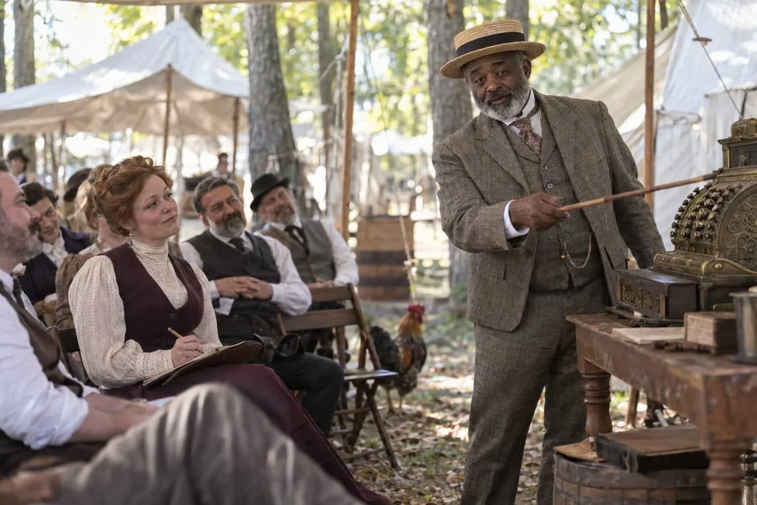 A reenactor teaches an 1890s-style sales class beside an antique cash register under camp tents.