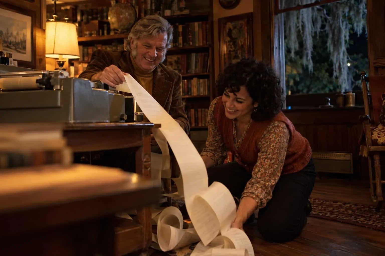 Two adults in 1970s clothes read a long paper printout spilling from a vintage teletype in a camp lodge.