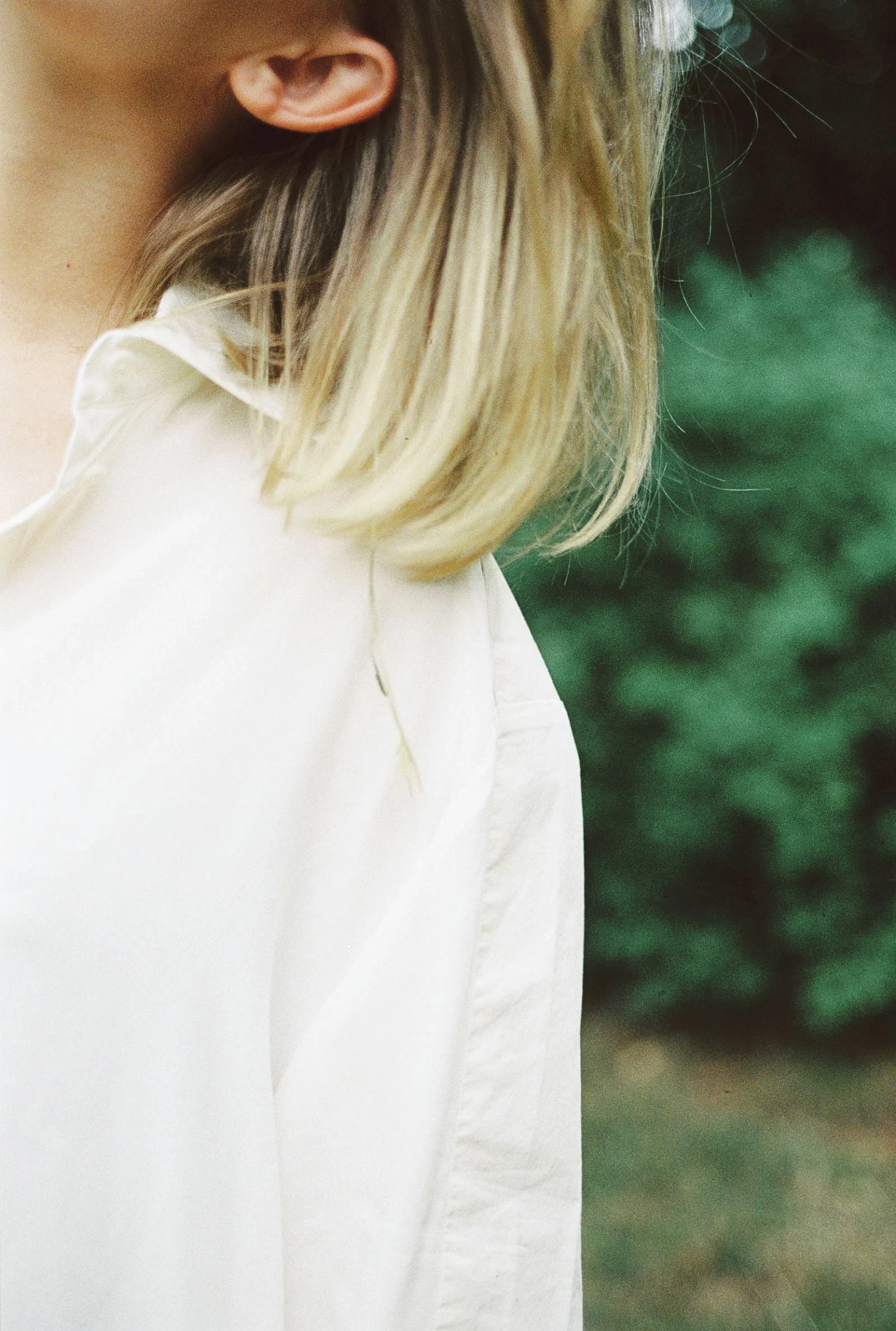 Close-up of a person with blonde hair wearing a white shirt, standing outdoors with green foliage in the background.