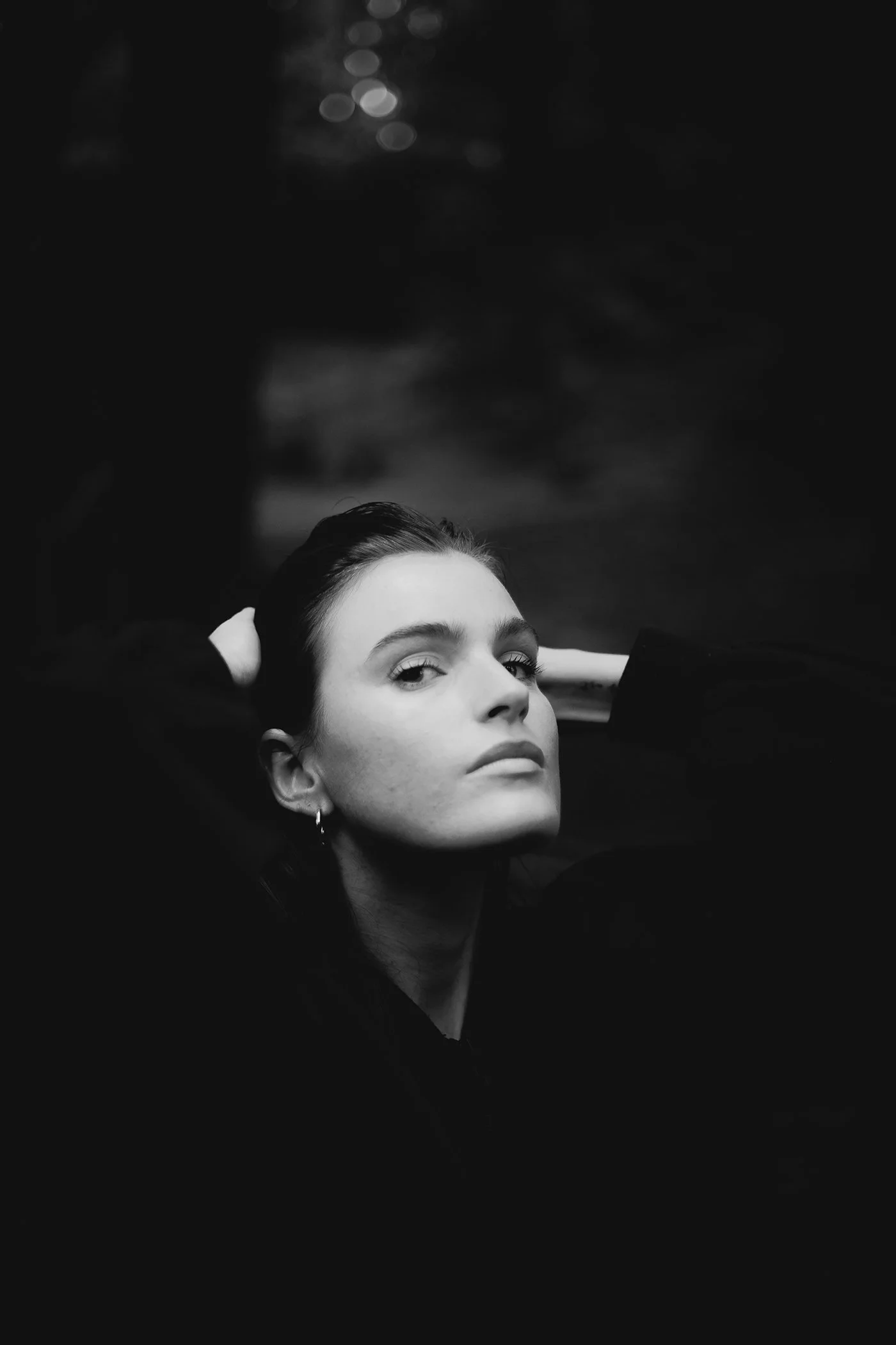 A black and white image of a woman with dark hair styled back, looking confidently at the camera with her hands behind her head, in a dark setting.