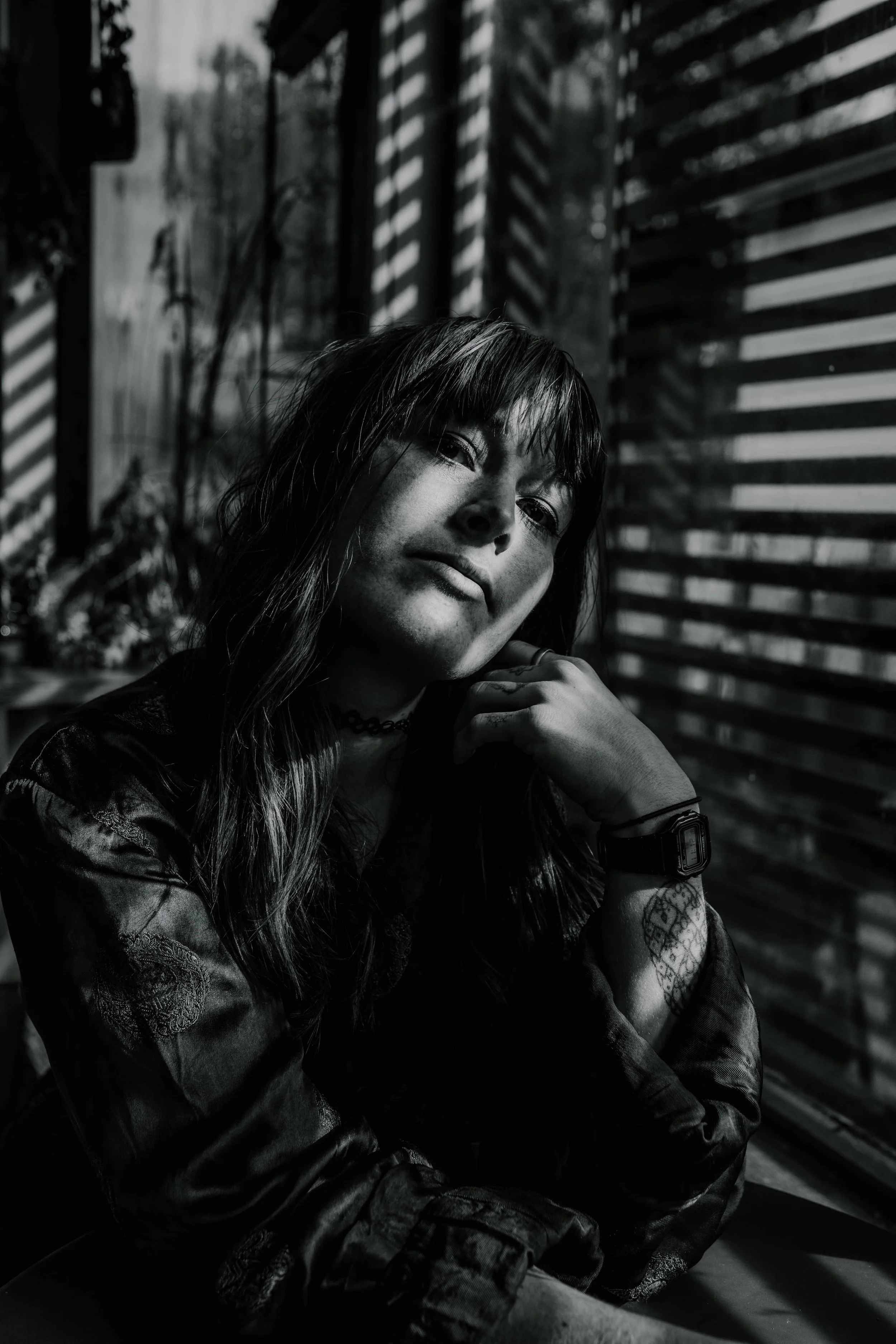 Black and white photo of a young woman with shoulder-length hair and a choker, sitting indoors next to a window with blinds, with natural light creating shadows on her face and background.