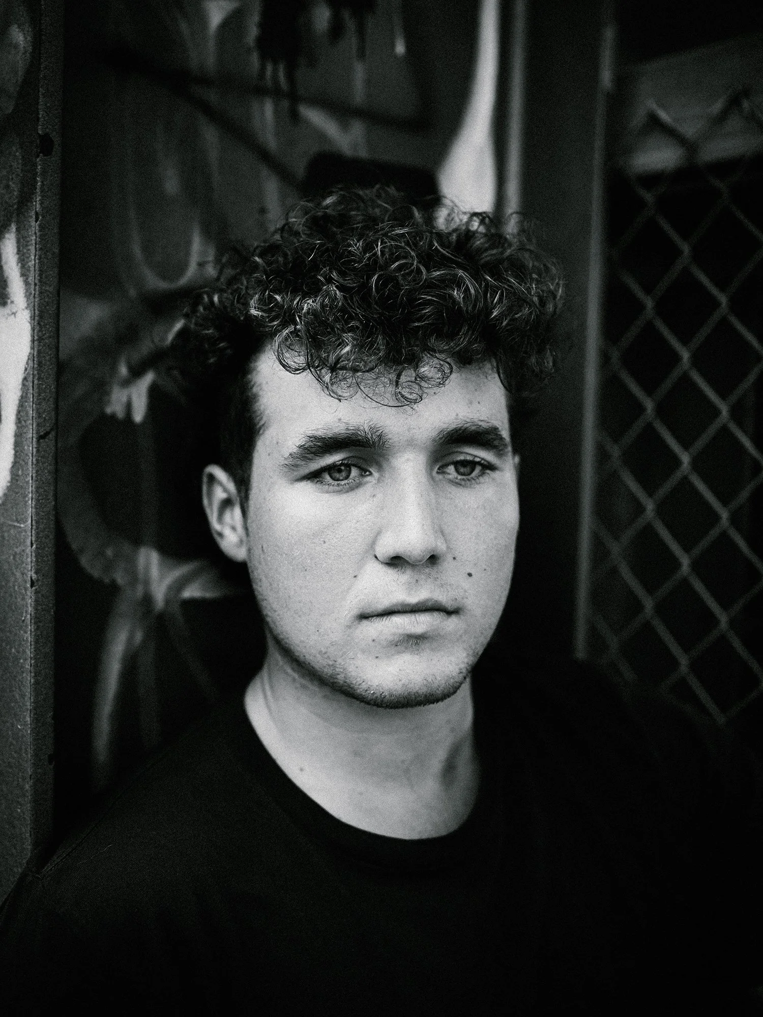 Black and white portrait of a young man with curly hair and a serious expression, sitting against a wall with a chain-link fence in the background.