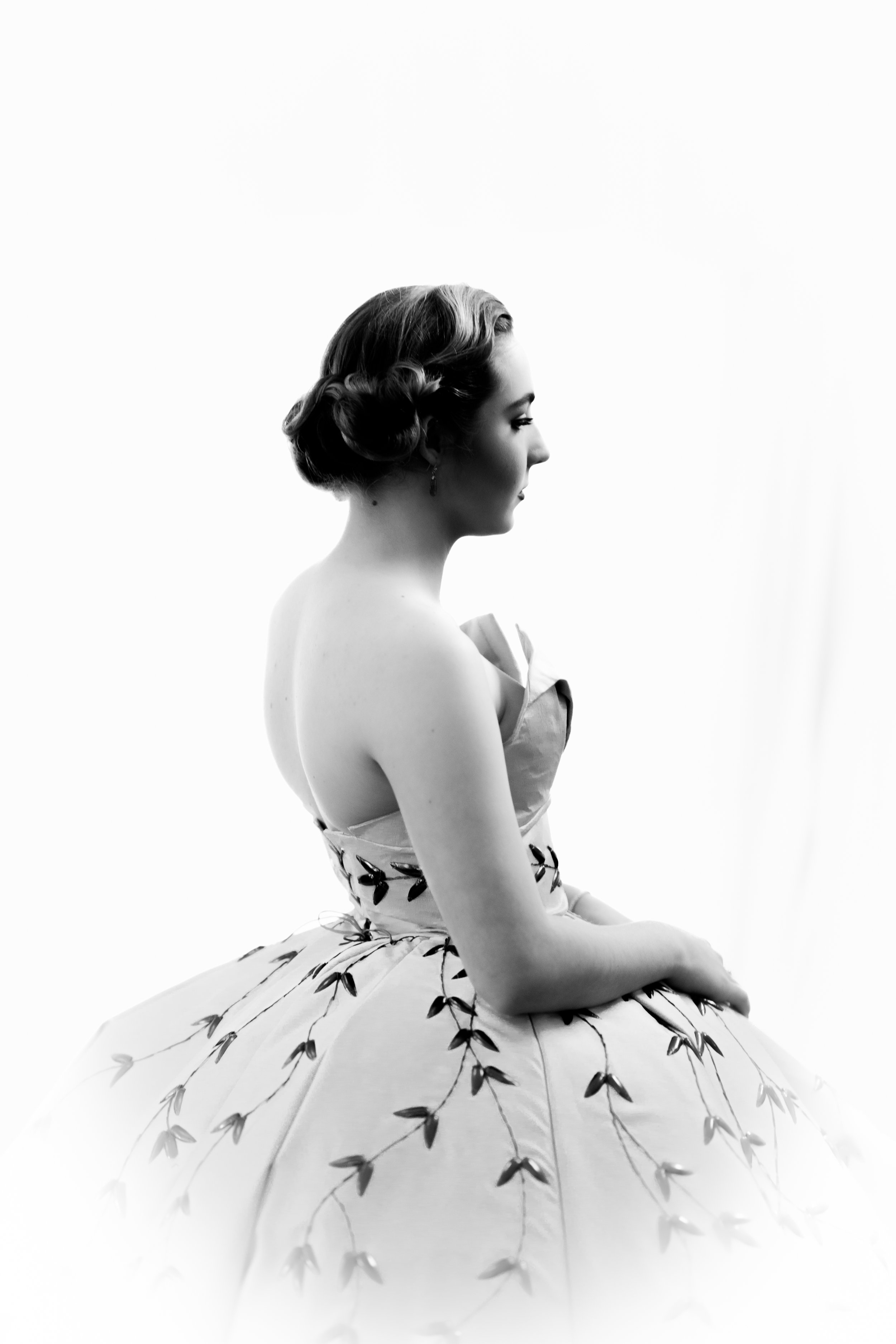 Black and white photo of a woman in a strapless, floral-embroidered gown, sitting with eyes closed, and her hair styled in vintage waves.