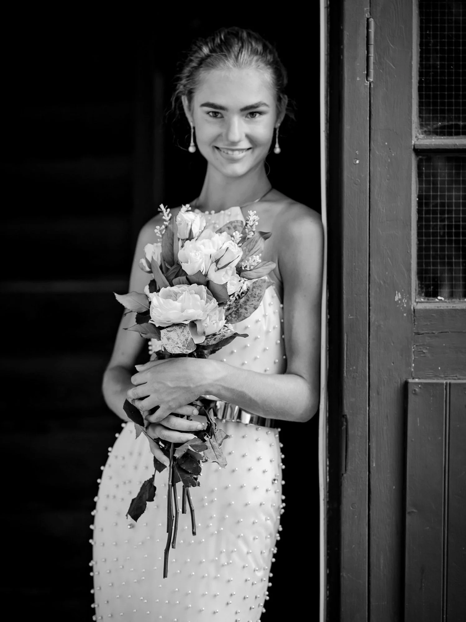 Black and white photo of a young woman holding a bouquet of flowers, wearing a dress decorated with small beads, standing at a doorway, smiling.