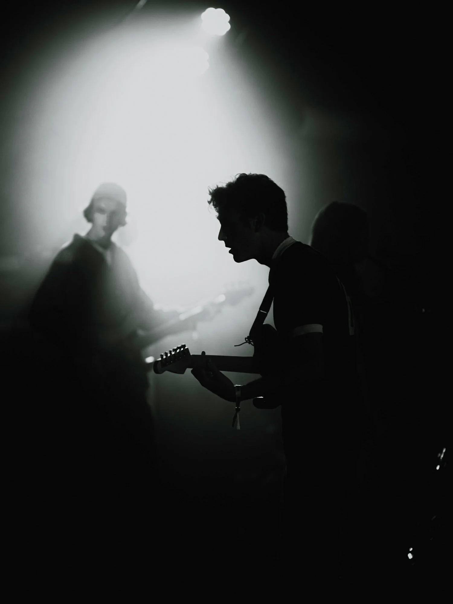 Silhouettes of musicians with guitars on stage in a dark, smoky setting, illuminated by a bright overhead light.