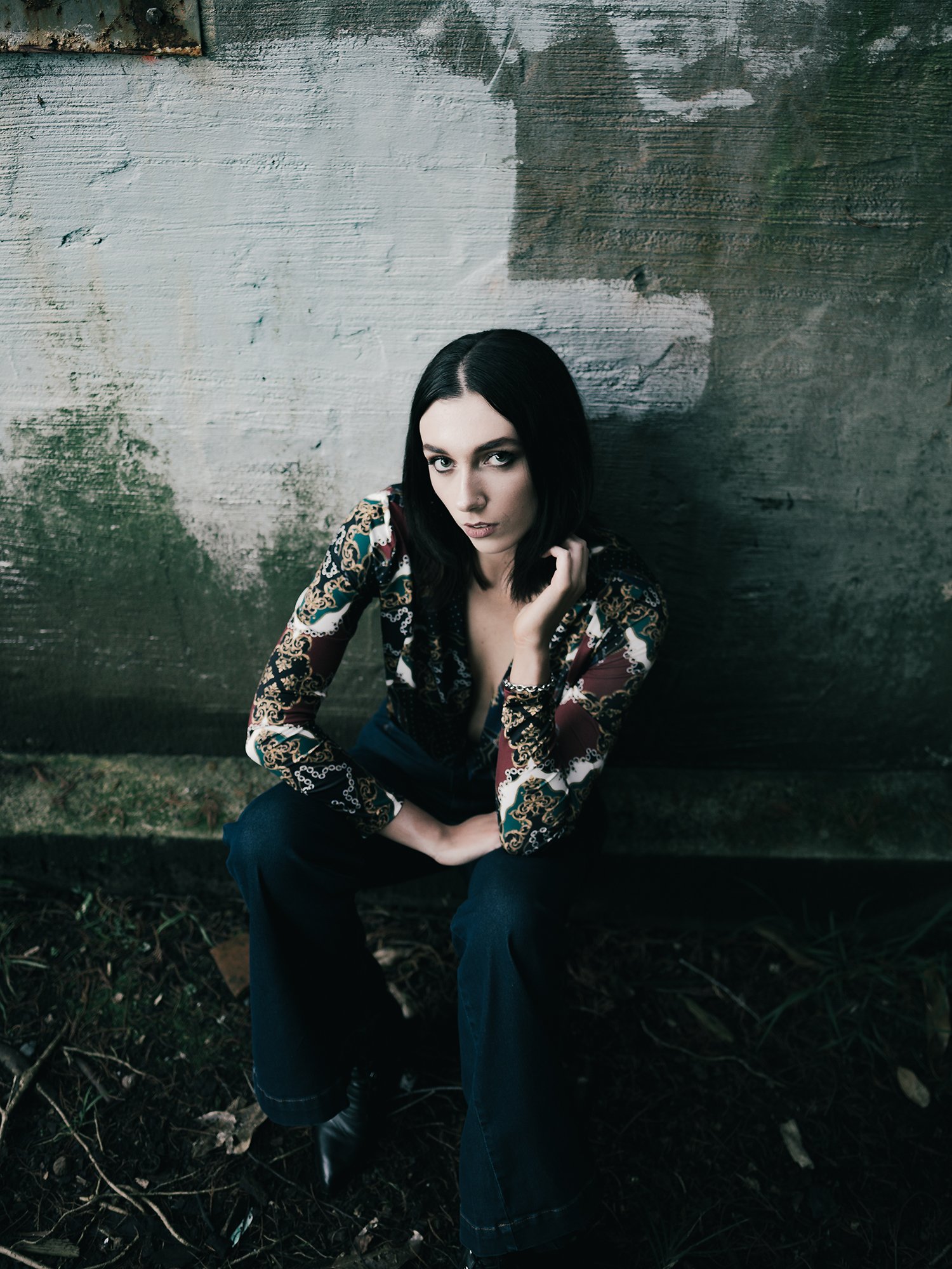 A woman with dark hair and a patterned blouse sitting on the ground against a weathered, textured wall outdoors.
