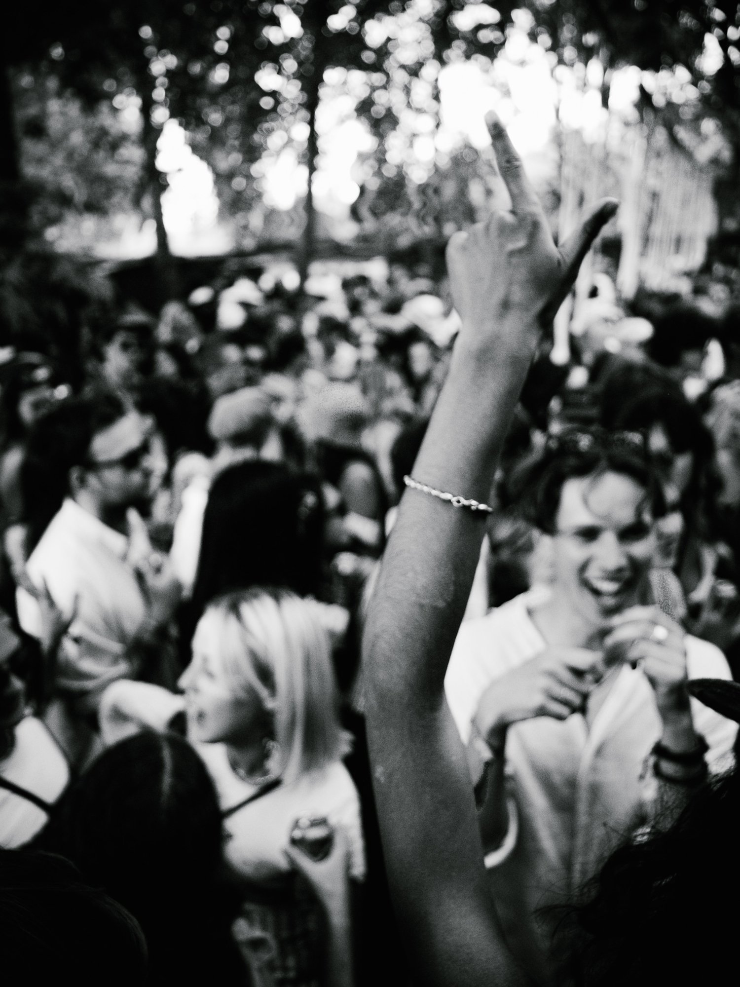 A black and white photo of a crowded outdoor event with people close together, one person raising their hand with a bracelet on their wrist, smiling woman holding a microphone, and trees in the background.