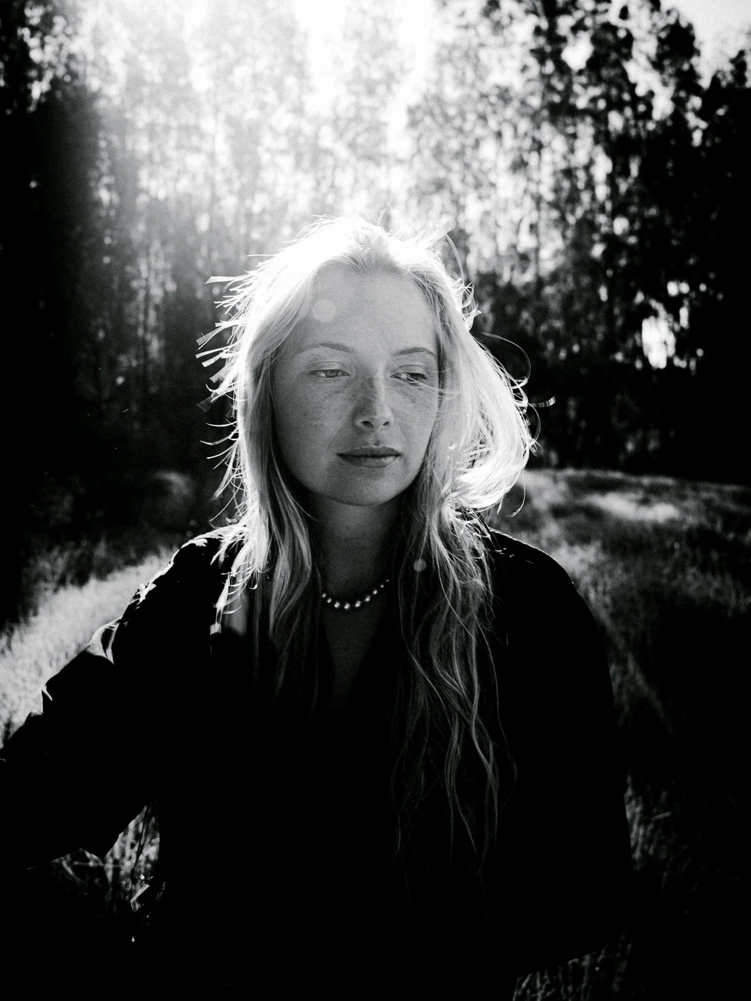 A woman with long blonde hair stands in a field as the sun shines behind her