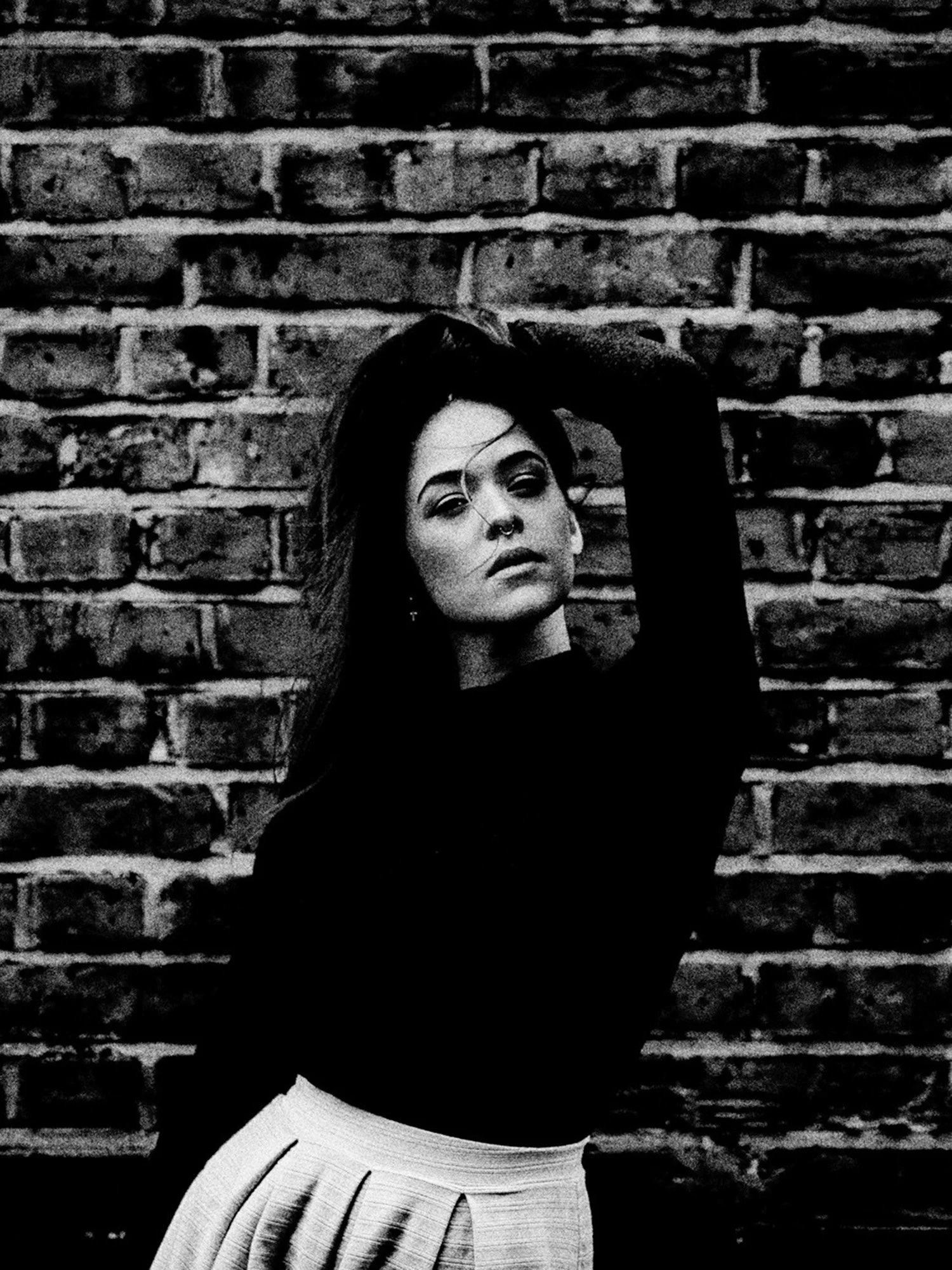 A woman with long hair and standing in front of brick wall raises her arm to her head