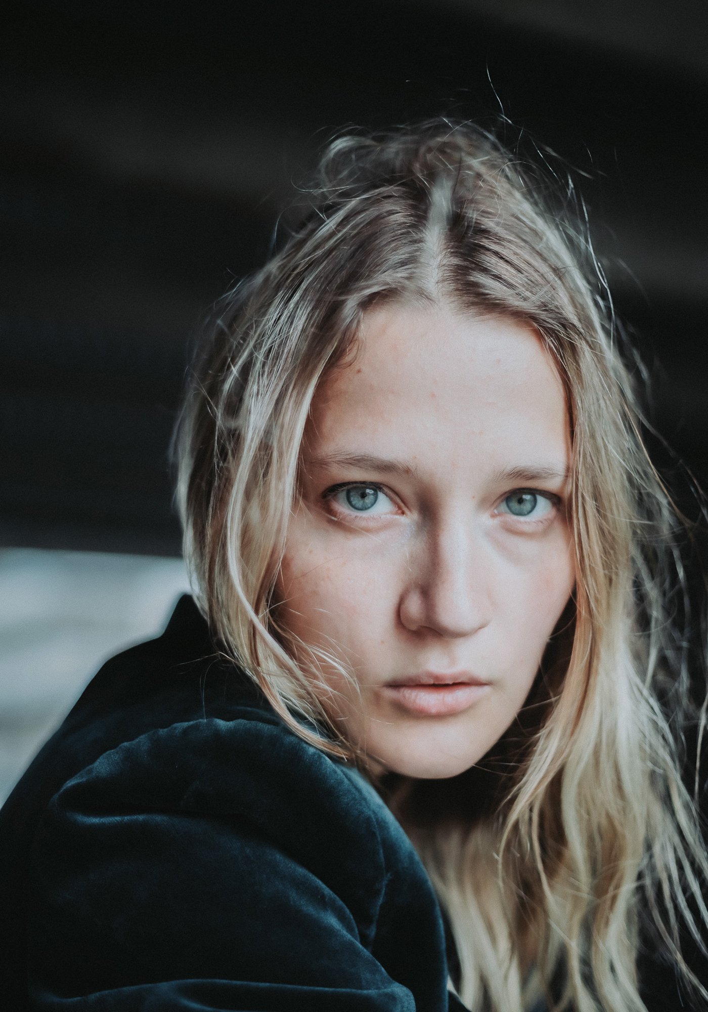 Close-up of a young woman with blonde hair and blue eyes, looking directly at the camera, with a dark background.