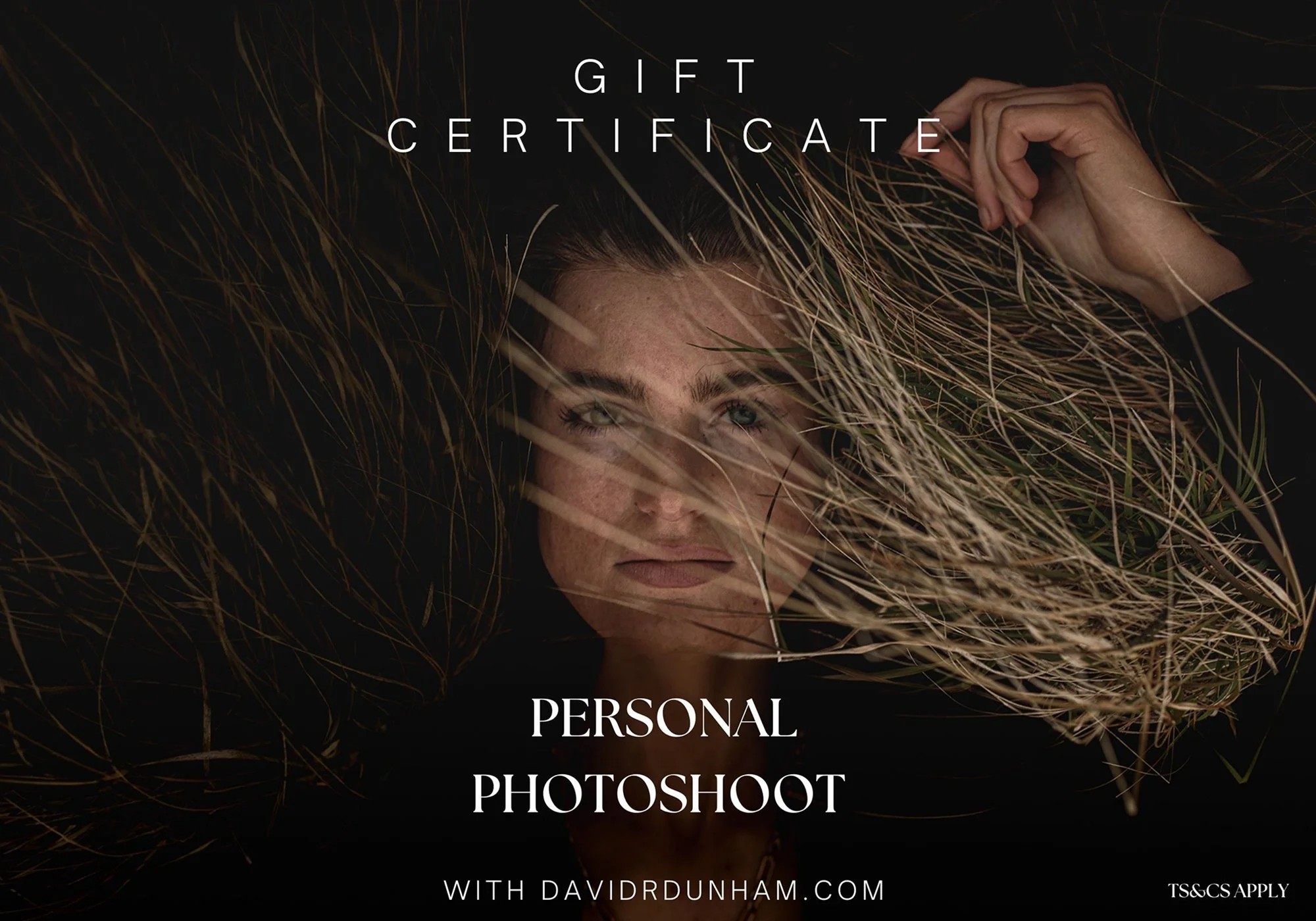 A woman lying on the ground with her face partially covered by dry grass, looking directly at the camera. The image is overlaid with text promoting a gift certificate for a personal photoshoot with davidrdunham.com.