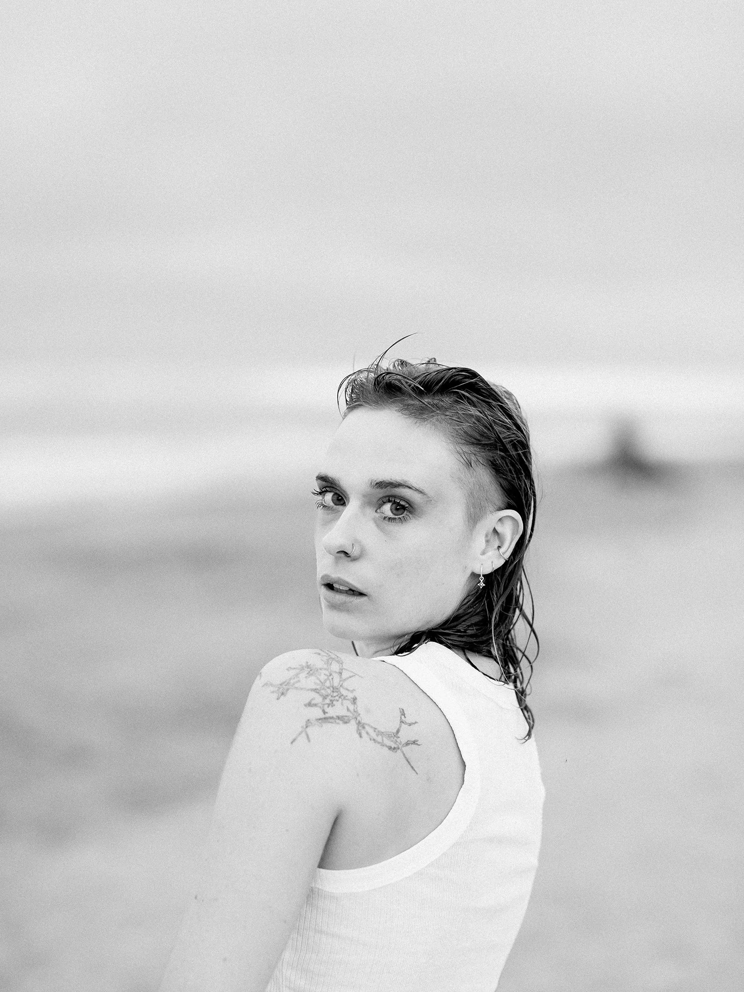 A black and white portrait of a young woman with wet hair, wearing a sleeveless top, looking over her shoulder at the camera, with a blurred beach or ocean background.