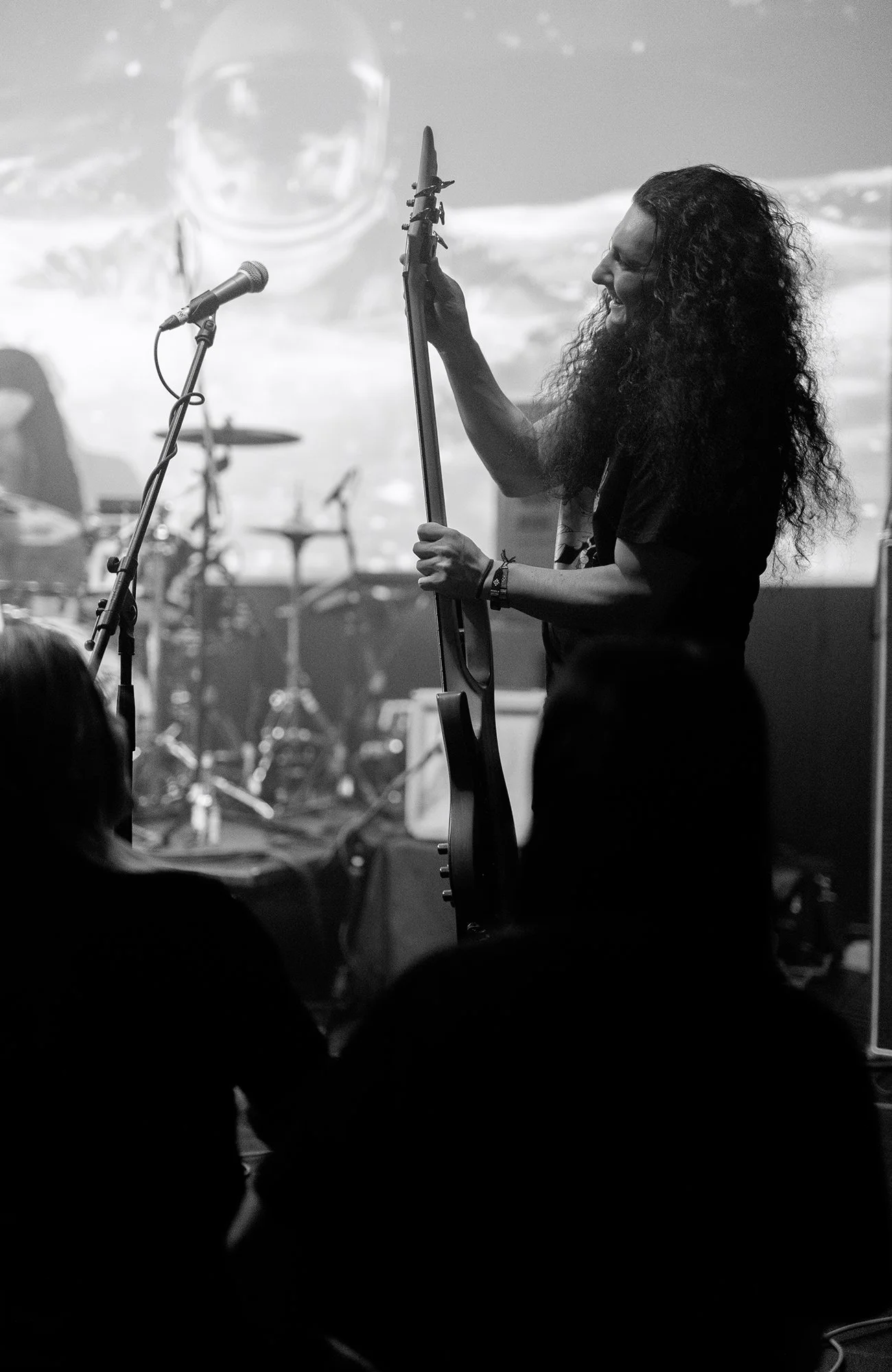 Black and white photo of a musician with long curly hair playing an electric guitar on stage with a drum set in the background.