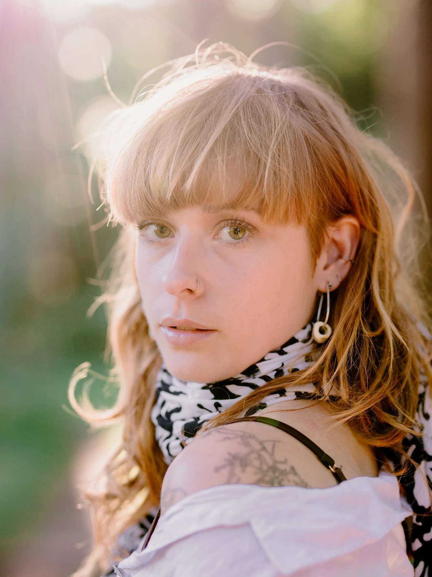 A young woman with light skin, green eyes, and red hair styled with bangs, wearing a black and white scarf, hoop earrings, and a white top with a tattoo on her shoulder, standing outdoors in sunlight.