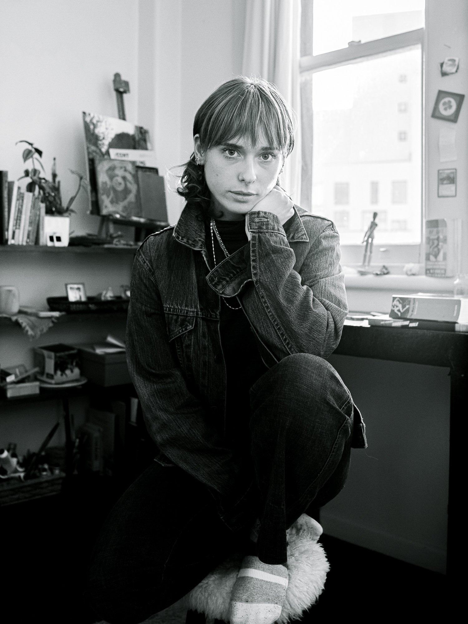 Black and white photo of a young woman with short hair, sitting on a furry stool in a cluttered room, looking directly at the camera with her hand resting on her chin, dressed in a denim jacket and striped socks.