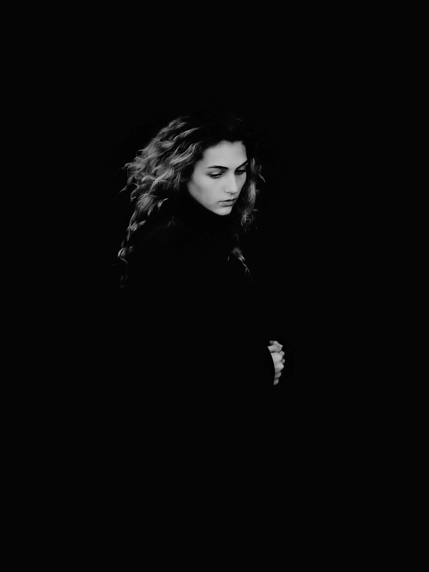 A young woman with curly hair looking downwards against a black background.